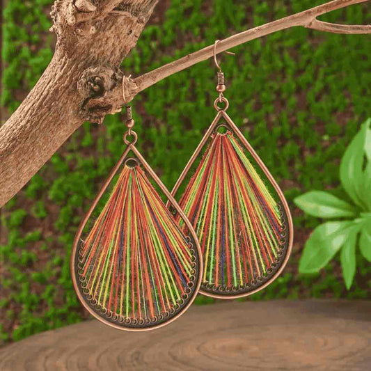 Close-up of a pair of multicolor threaded boho earrings hanging from a tree branch, with people blurred in the background.