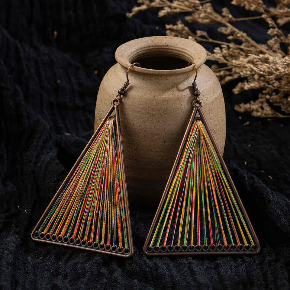 Pair of boho earrings with bronze triangle frames and multicolor threaded string art, displayed against a dark fabric and rustic pot.