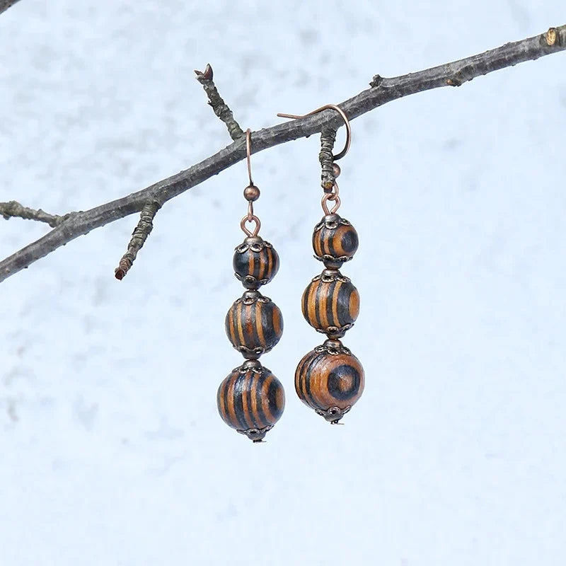 A pair of boho wooden beaded drop earrings with black and brown stripes and swirls, hanging from a bare tree branch against a light background.