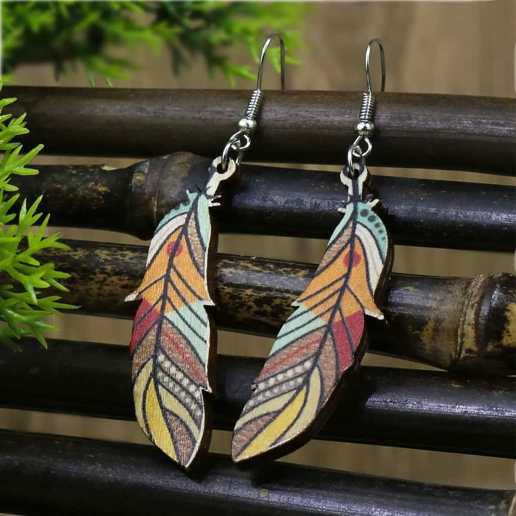 Close-up of a pair of boho wooden feather earrings with a colorful tribal pattern, hanging from silver hooks against a dark wooden background with green foliage.