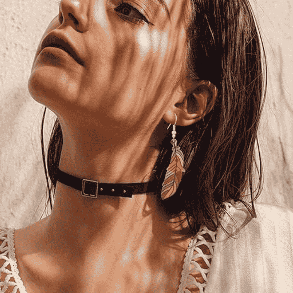 A woman wearing a single boho wooden feather earring, looking upwards, with a black choker necklace. The earring has a colorful tribal pattern.