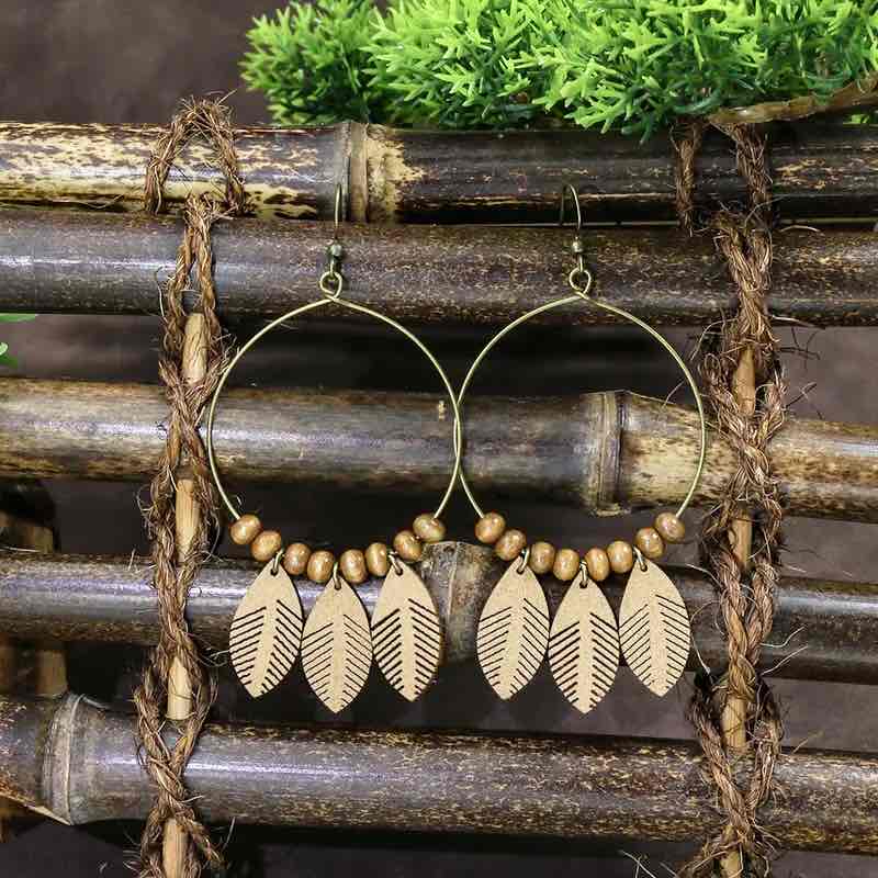 A pair of large boho earrings with wooden leaf charms and brown beads on a gold-toned hoop, displayed against a rustic bamboo background with green foliage.