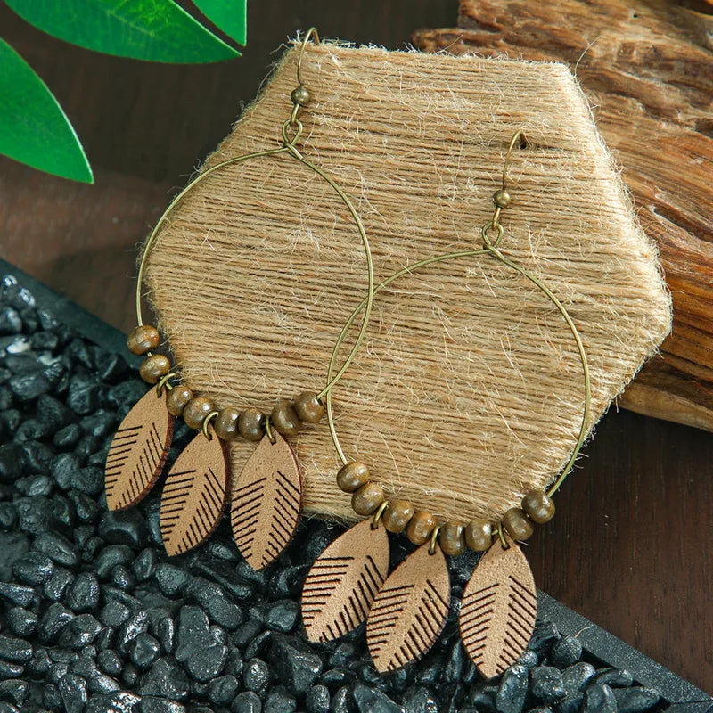 A single boho earring featuring a gold-toned hoop, brown wooden beads, and three wooden leaf charms with etched details, resting on a textured jute surface with dark pebbles.