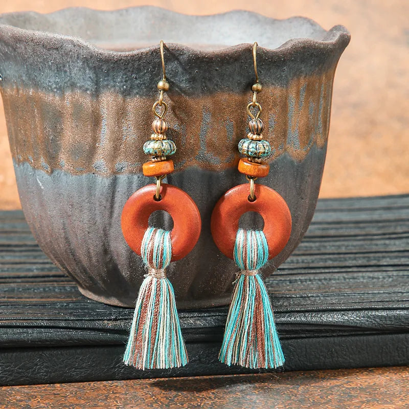 A pair of boho earrings featuring wooden hoops and blue, brown, and white tassels, displayed in a rustic ceramic bowl.