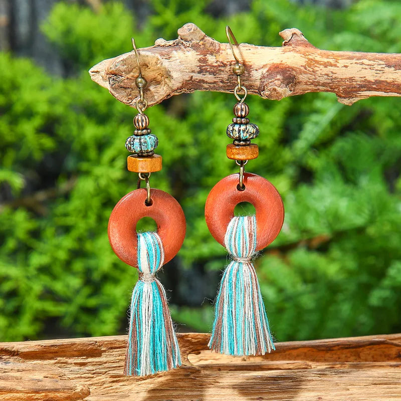 A pair of boho earrings with wooden hoops and blue, brown, and white tassels, hanging from a natural tree branch against a green foliage background.