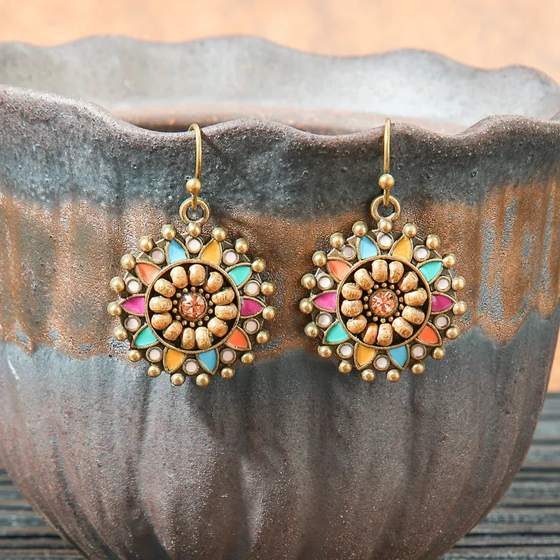 Two boho earrings with a round, floral design, wooden beads, multicolor enamel, and a central stone, hanging against a rustic ceramic pot.
