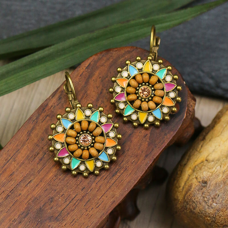 A pair of boho earrings with a round, floral design, wooden beads, multicolor enamel, and a central stone, resting on a carved wooden surface.