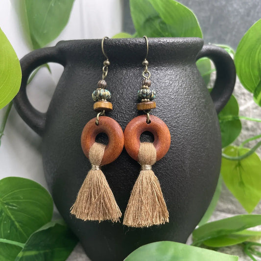 A pair of boho wooden tassel earrings with earthy rings and beaded accents, hanging against a dark pot with green foliage.