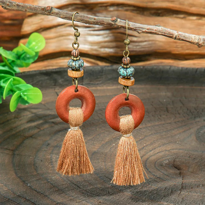 A close-up of boho wooden tassel earrings with wooden rings and beaded details, resting on a rustic wooden surface.