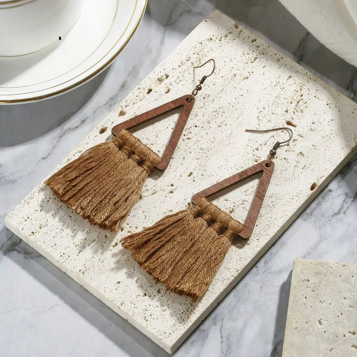 A pair of brown boho earrings with wooden triangles and macrame tassels, displayed on a white marble surface.