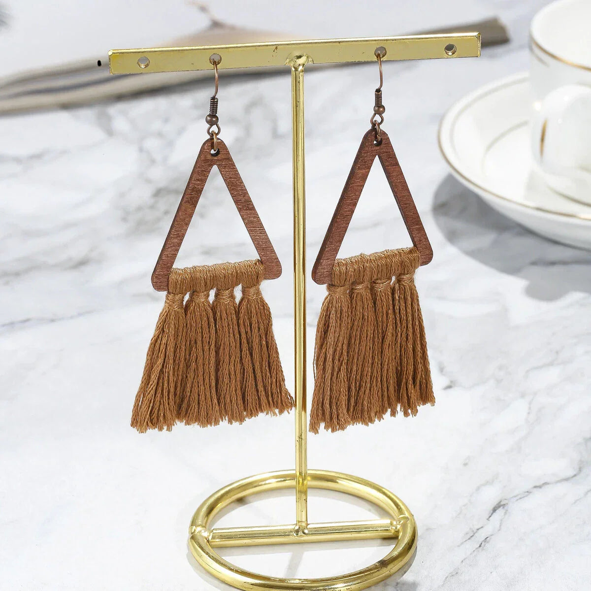 A pair of brown boho earrings with wooden triangles and macrame tassels, hanging on a golden earring stand against a marble background.