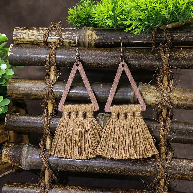 A pair of brown boho earrings with wooden triangles and macrame tassels, displayed on a rustic bamboo surface with green foliage in the background.