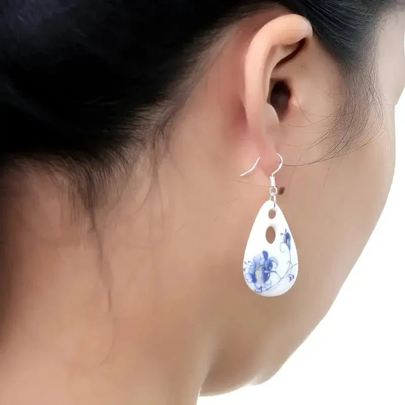A woman wearing a single white ceramic drop earring with a blue floral pattern, showcasing its size and how it hangs.