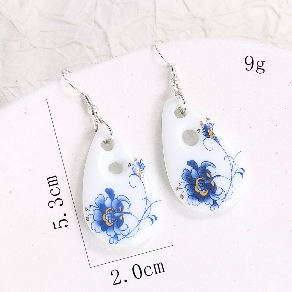 A pair of white ceramic drop earrings with blue floral designs, showing their dimensions (5.3cm length, 2.0cm width) and weight (9g).