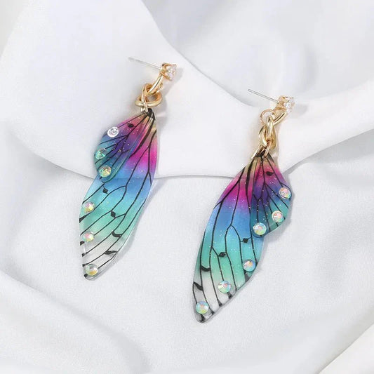 A pair of colorful acrylic butterfly long earrings with blue, green, and pink gradient wings, adorned with small clear rhinestones, displayed on a white draped fabric.