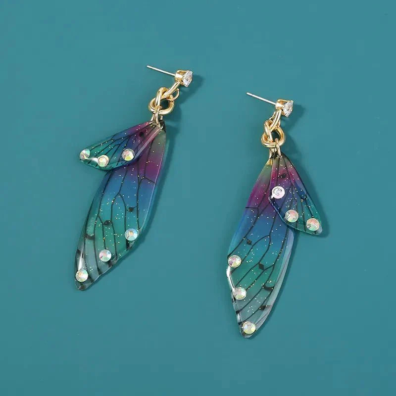 A pair of colorful acrylic butterfly long earrings featuring blue, green, and purple gradient wings with black vein details and scattered clear rhinestones, presented on a teal background.