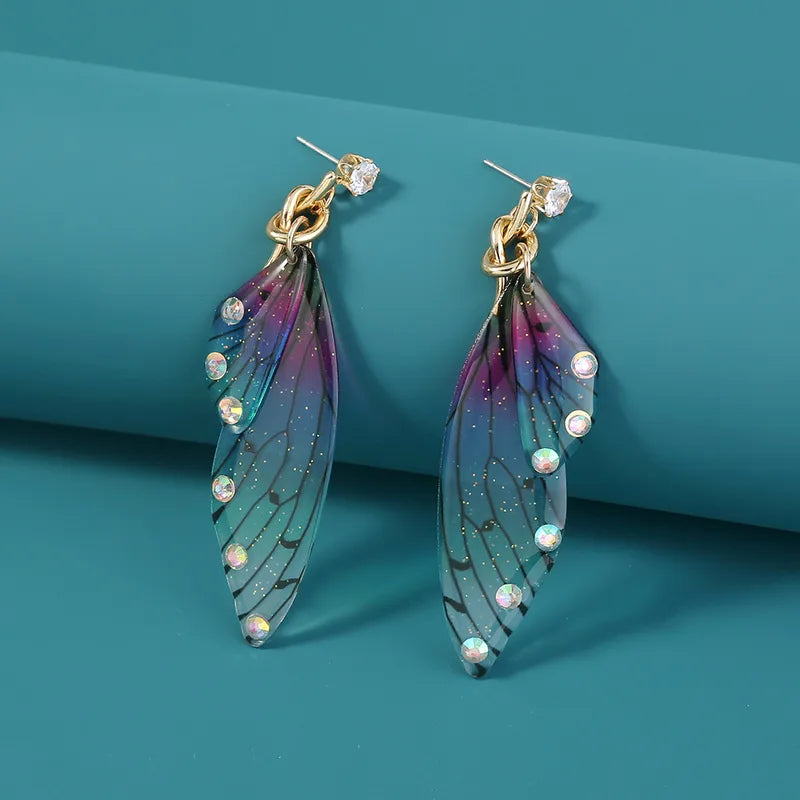A pair of vibrant acrylic butterfly long earrings showcasing blue, purple, and green gradient wings with black vein patterns and clear rhinestone accents, set against a teal background.