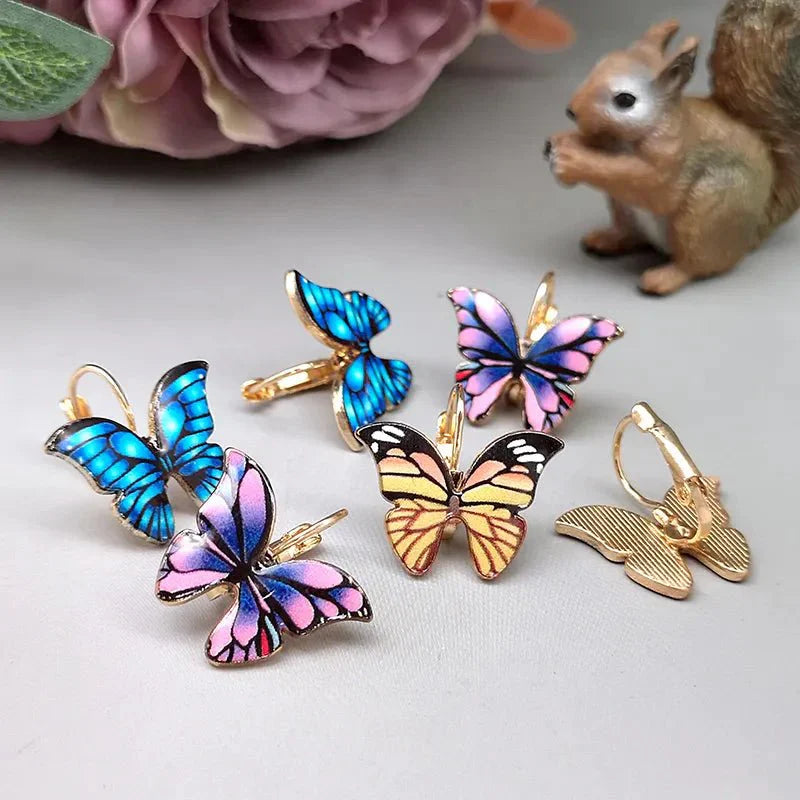 A collection of colorful enamel butterfly korean earrings in various shades (blue, pink, yellow) displayed on a light surface with a squirrel figurine in the background.