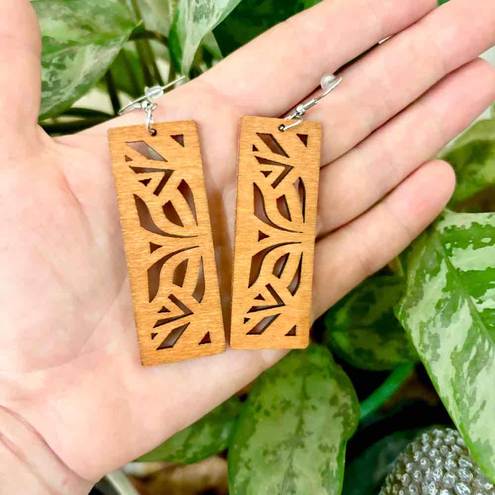 A hand holding a pair of light brown wooden earrings with leaf cutouts, set against a backdrop of green foliage, highlighting the natural aesthetic of the wooden earrings.