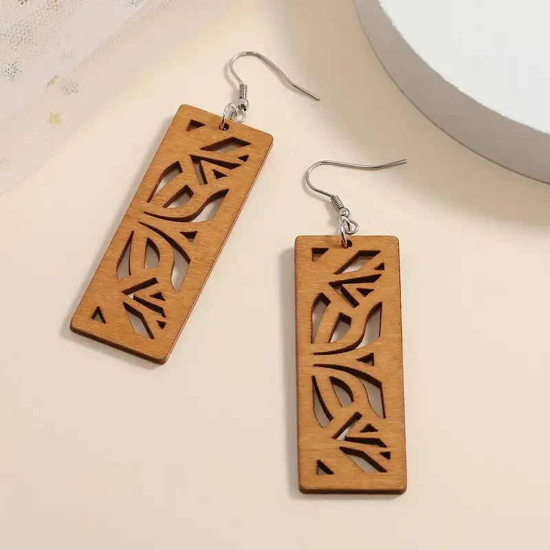 A pair of light brown wooden earrings with detailed leaf cutouts, showcasing their rectangular drop design and silver-toned fish hooks, on a light surface.