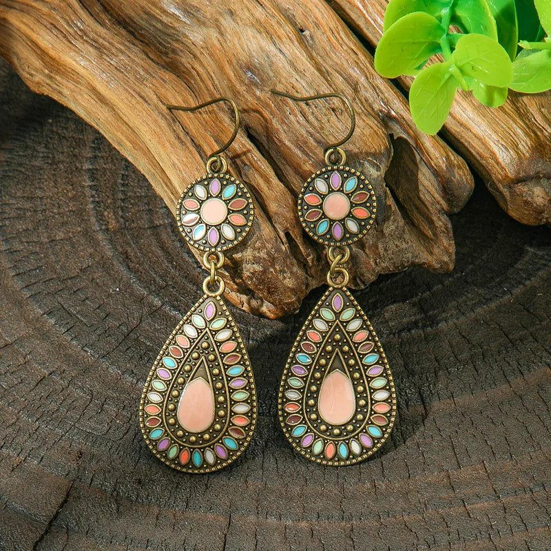 A pair of oxidised earrings with multi-color enamel floral and teardrop designs, resting on a wooden surface with green leaves in the background.