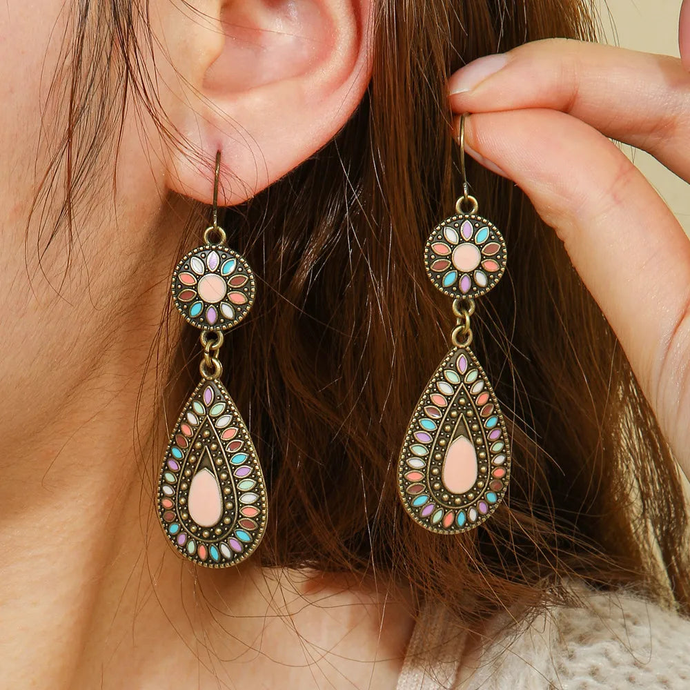 A woman wearing a pair of oxidised earrings with multi-color enamel floral and teardrop designs, with her hand gently touching one earring.