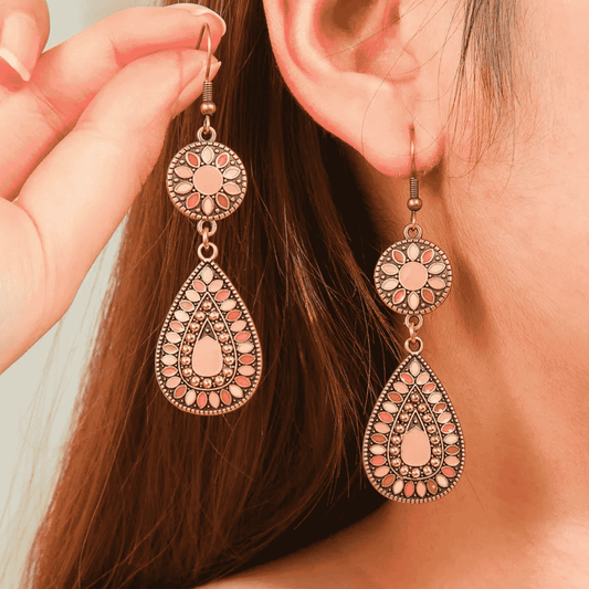 A woman wearing a pair of long oxidised earrings with floral and teardrop designs in pink and white enamel.