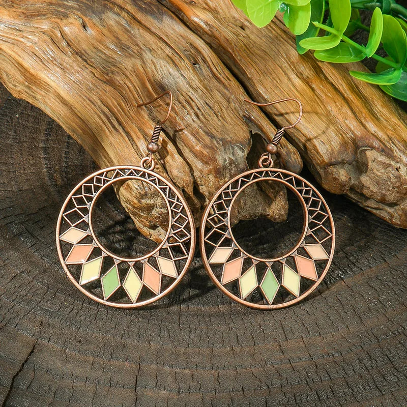 A pair of oxidised earrings with a geometric hoop design and pastel enamel diamonds, displayed on a wooden surface with green foliage.