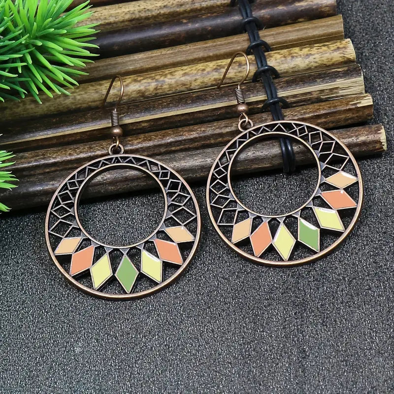 Two oxidised earrings, featuring a geometric hoop pattern with colorful enamel diamonds in pastel shades, set against a dark background with bamboo sticks and green leaves.