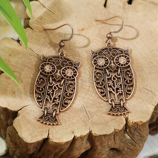 A pair of oxidised earrings shaped like owls, featuring a filigree design with small rhinestones for eyes, displayed on a wooden surface.