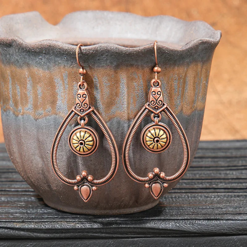 A pair of oxidised teardrop drop earrings with a sunburst and filigree design, displayed in a rustic brown ceramic pot.