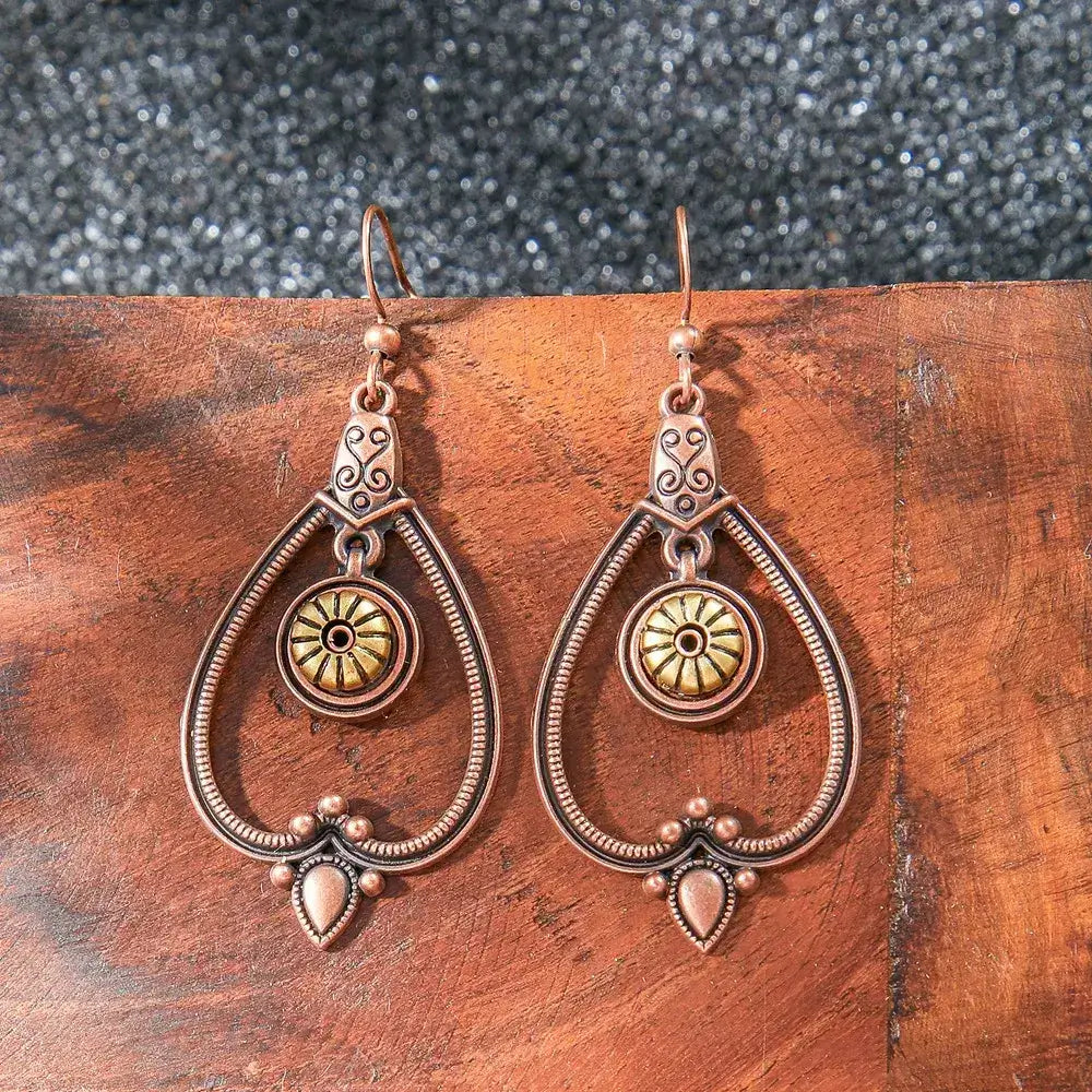A pair of oxidised teardrop drop earrings featuring a sunburst and filigree pattern, resting on a wooden surface.