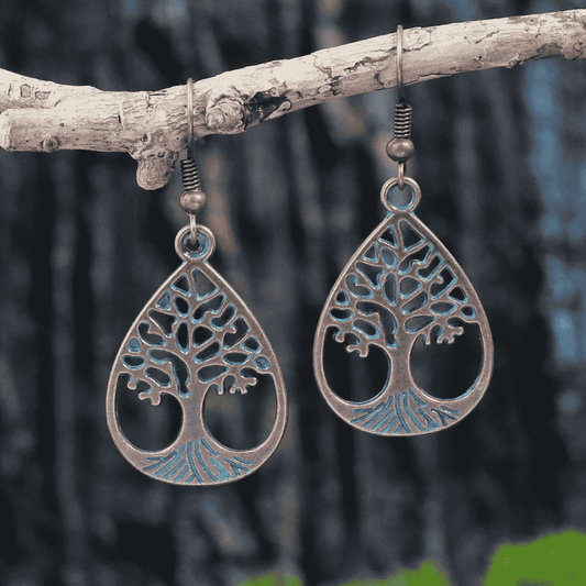 Pair of oxidised earrings, teardrop shape with a Tree of Life design and blue accents, hanging from a light-colored tree branch, against a blurred dark background. These are oxidised earrings.