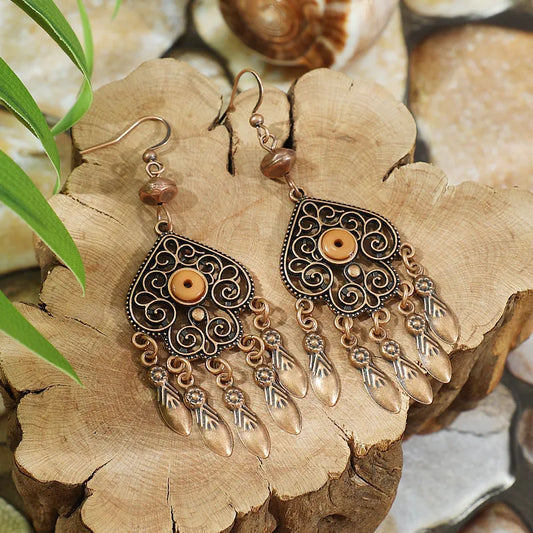 A pair of oxidised earrings with antique copper filigree and tan bead accents, displayed on a rustic wooden slab with green leaves and seashells in the background.