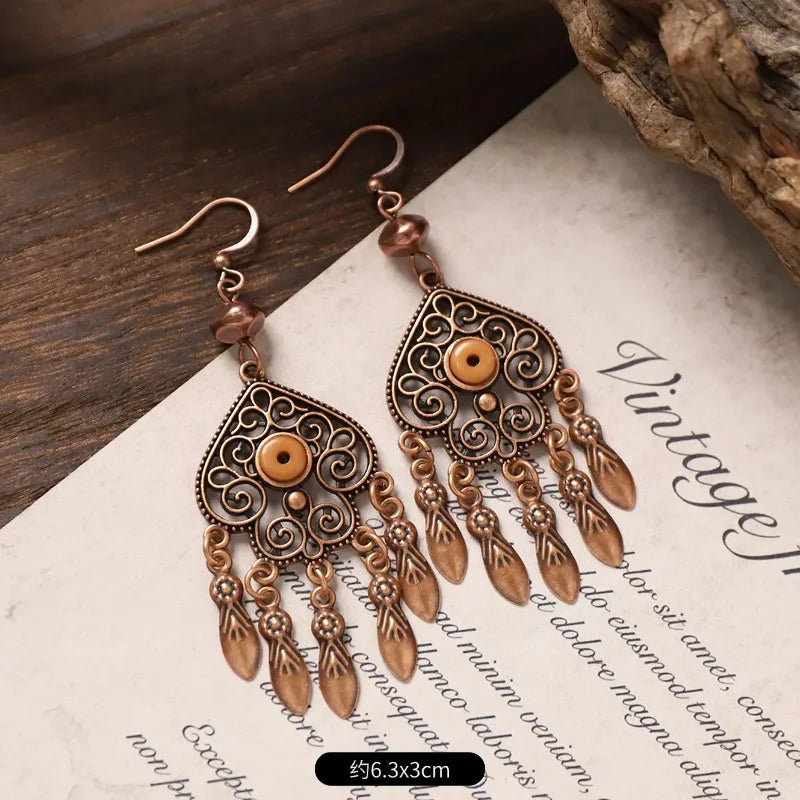 A pair of oxidised earrings with antique copper filigree and tan bead accents, laid on a vintage-style paper with text and a piece of wood, showing the approximate size of 6.3x3cm.
