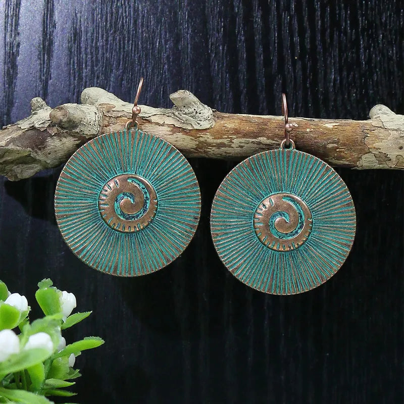 A pair of oxidised blue boho earrings with a spiral disc design, hanging from a wooden branch.