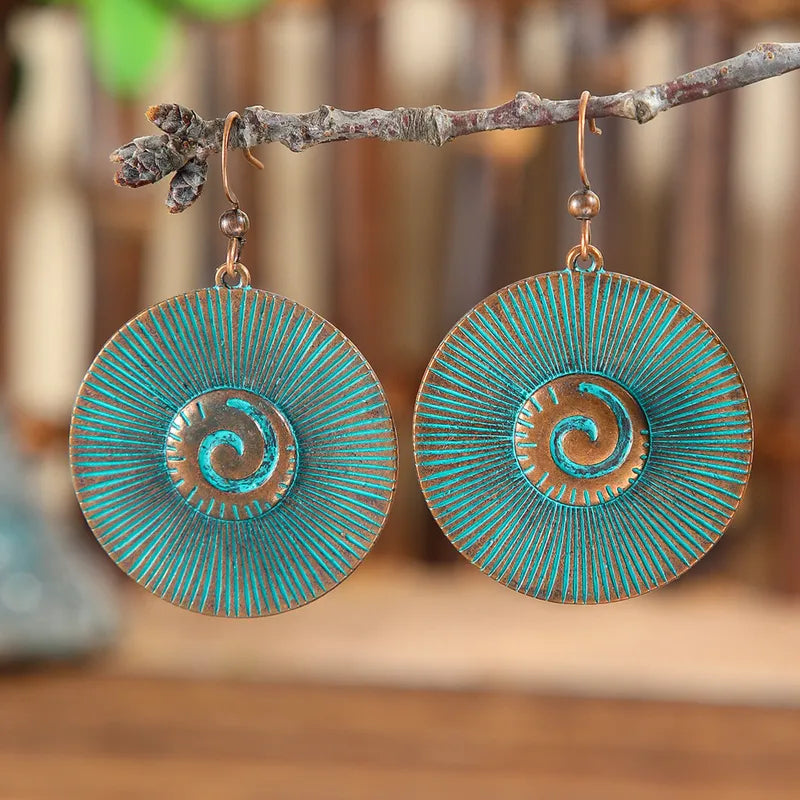 Close-up of a pair of oxidised blue boho earrings with a spiral disc design, hanging from a thin branch.
