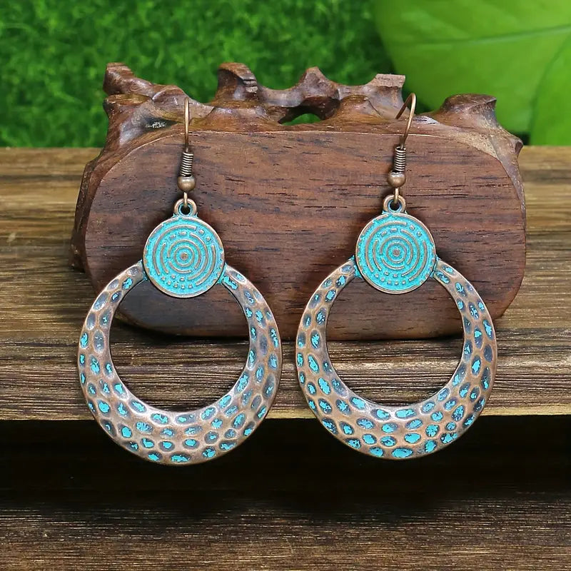 A pair of oxidised earrings with turquoise blue patina and a spiral disc design, resting on a wooden block.