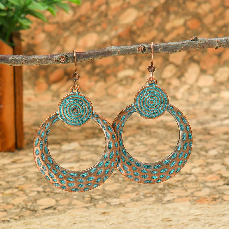 A pair of oxidised earrings with turquoise blue patina and a spiral disc design, hanging from a tree branch.