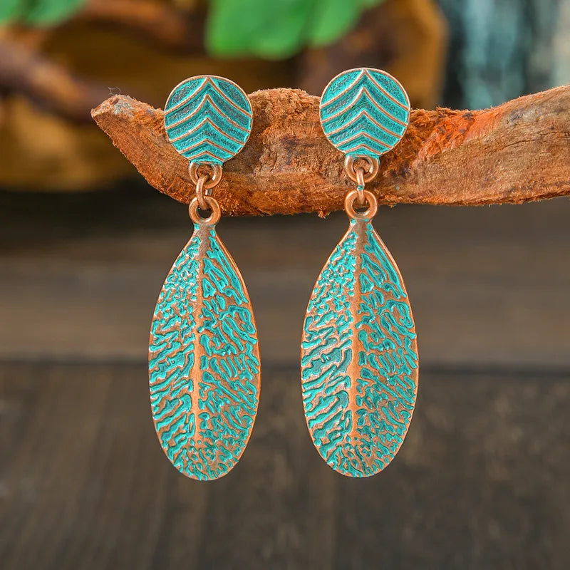A pair of oxidised earrings with copper-toned leaf drops and turquoise blue patina, featuring chevron-patterned studs, displayed on a wooden branch.