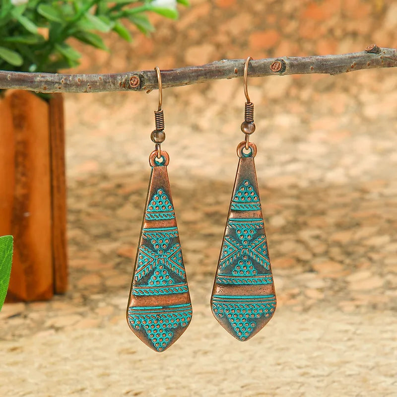 A pair of oxidised earrings with blue tribal and geometric patterns, shaped like elongated teardrops, hanging from a branch.
