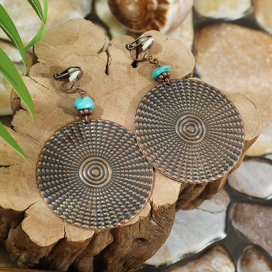 A pair of large, round oxidised earrings with a textured, geometric pattern and turquoise beads, displayed on a wooden surface. These oxidised earrings feature clip-on backs.