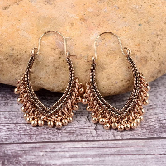 A pair of oxidised earrings with a ghungroo jingle design, featuring an antique bronze finish and black accents, displayed on a rustic stone and wooden surface.