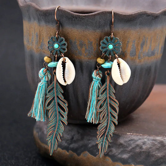 Close-up of oxidised unique boho dangle earrings with shell and feather charms, featuring turquoise beads and tassels, set against a dark ceramic pot.