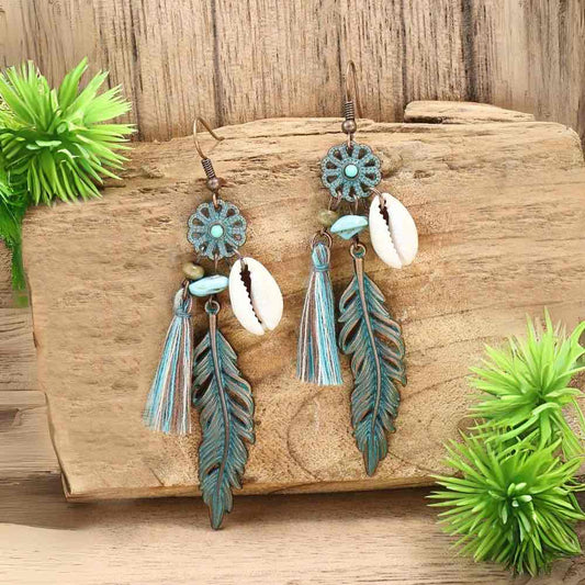 Oxidised unique boho dangle earrings with shell and feather charms, turquoise accents, and tassels, displayed on a rustic wooden surface with green plants.