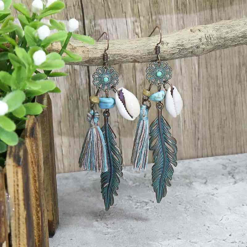 Oxidised unique boho dangle earrings with shell and feather charms, turquoise beads, and tassels, hanging from a tree branch with green foliage in the background.
