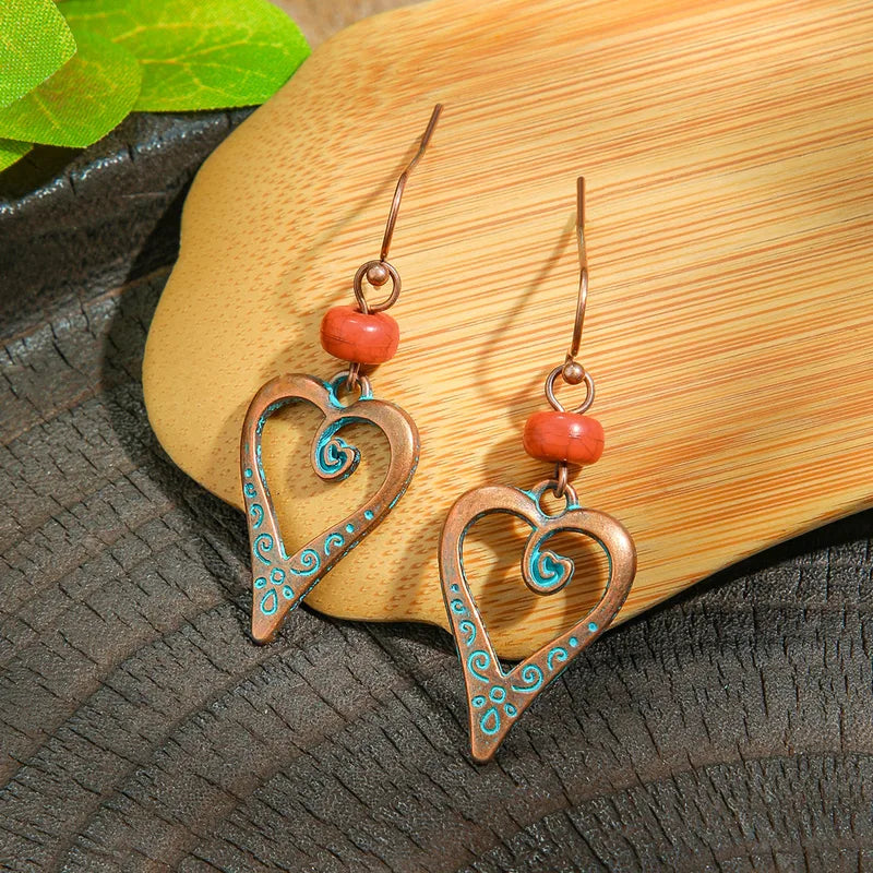 A pair of oxidised boho heart drop earrings with teal swirls and red beads, resting on a wooden surface.