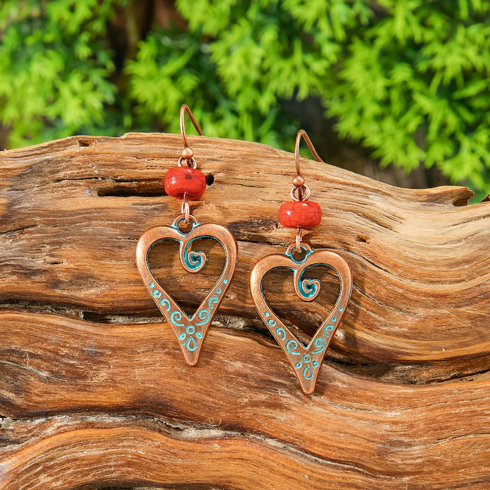 A pair of oxidised boho heart drop earrings with teal swirls and red beads, displayed on a natural wooden log.