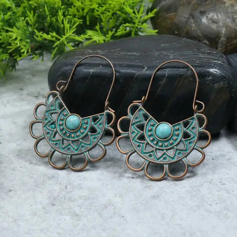 A pair of oxidised earrings with a mandala design and a turquoise stone, resting on a grey surface with green foliage and dark rocks in the background, highlighting the oxidised earrings.