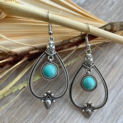 A pair of oxidised boho earrings with a teardrop shape and turquoise stone, displayed against dried leaves and wood.